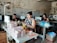 A group of people work at sewing machines in a workshop with worn and peeling walls. The workspace is filled with stacks of fabric pieces, spools of thread, and sewing equipment. The individuals are focused on their tasks, handling different materials.