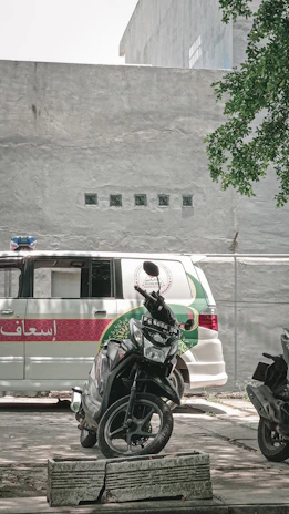 A scooter is parked in front of an ambulance with a white and green design. The scene takes place in an urban area with a concrete wall and some greenery in the background.