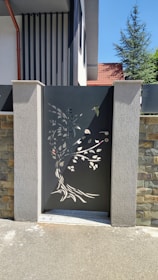 A decorative metal gate with intricate cutouts depicting a tree and leaves is set between two concrete pillars. The gate features a dark color, contrasting with the stone and concrete textures surrounding it. The background includes part of a residential building with a slanted red roof and a tree visible over the wall.