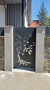A decorative metal gate with intricate cutouts depicting a tree and leaves is set between two concrete pillars. The gate features a dark color, contrasting with the stone and concrete textures surrounding it. The background includes part of a residential building with a slanted red roof and a tree visible over the wall.