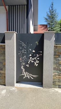 A decorative metal gate with intricate cutouts depicting a tree and leaves is set between two concrete pillars. The gate features a dark color, contrasting with the stone and concrete textures surrounding it. The background includes part of a residential building with a slanted red roof and a tree visible over the wall.