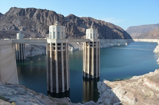 A large concrete dam structure spans across a river with cylindrical towers rising from the water. The surrounding landscape features rugged, rocky mountains under a clear blue sky.