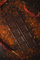 A dark chocolate bar is placed on a surface sprinkled with red pepper flakes. Two dried red chili peppers are positioned on either side of the chocolate, adding a spicy aesthetic to the arrangement.