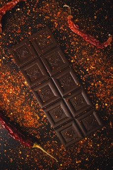 A dark chocolate bar is placed on a surface sprinkled with red pepper flakes. Two dried red chili peppers are positioned on either side of the chocolate, adding a spicy aesthetic to the arrangement.