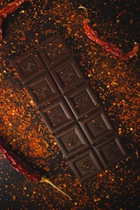 A dark chocolate bar is placed on a surface sprinkled with red pepper flakes. Two dried red chili peppers are positioned on either side of the chocolate, adding a spicy aesthetic to the arrangement.