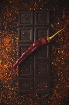 A dark chocolate bar is placed vertically on a surface scattered with red chili flakes. A single dried red chili pepper is laid diagonally across the chocolate, adding a pop of color and contrast to the dark, rich tones of the chocolate.