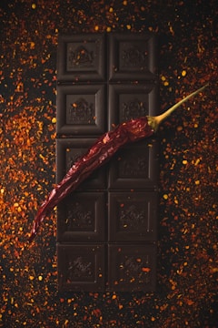 A dark chocolate bar is placed vertically on a surface scattered with red chili flakes. A single dried red chili pepper is laid diagonally across the chocolate, adding a pop of color and contrast to the dark, rich tones of the chocolate.