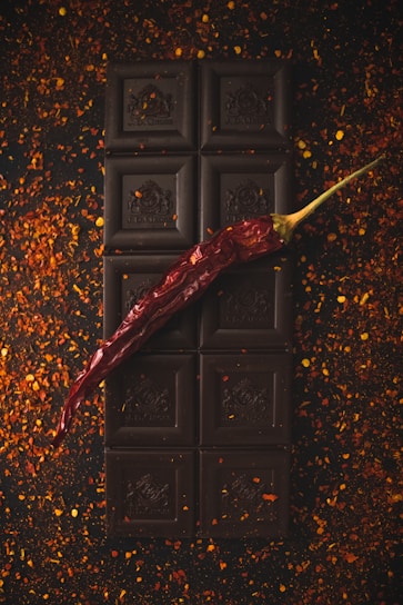 A dark chocolate bar is placed vertically on a surface scattered with red chili flakes. A single dried red chili pepper is laid diagonally across the chocolate, adding a pop of color and contrast to the dark, rich tones of the chocolate.