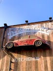 A red vintage car is affixed to a wooden wall, seemingly defying gravity. The wall bears the words 'Dependable' and 'Auto Repair' in large white letters. The car is an old model and has the phrase 'Mechanic on Duty' written on its side.