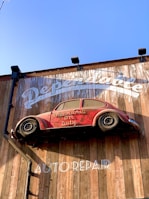 A red vintage car is affixed to a wooden wall, seemingly defying gravity. The wall bears the words 'Dependable' and 'Auto Repair' in large white letters. The car is an old model and has the phrase 'Mechanic on Duty' written on its side.