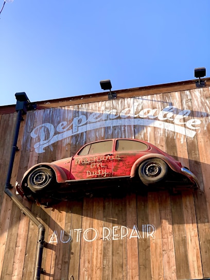 A red vintage car is affixed to a wooden wall, seemingly defying gravity. The wall bears the words 'Dependable' and 'Auto Repair' in large white letters. The car is an old model and has the phrase 'Mechanic on Duty' written on its side.