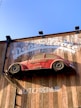 A red vintage car is affixed to a wooden wall, seemingly defying gravity. The wall bears the words 'Dependable' and 'Auto Repair' in large white letters. The car is an old model and has the phrase 'Mechanic on Duty' written on its side.