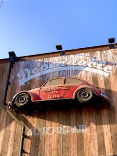 A red vintage car is affixed to a wooden wall, seemingly defying gravity. The wall bears the words 'Dependable' and 'Auto Repair' in large white letters. The car is an old model and has the phrase 'Mechanic on Duty' written on its side.