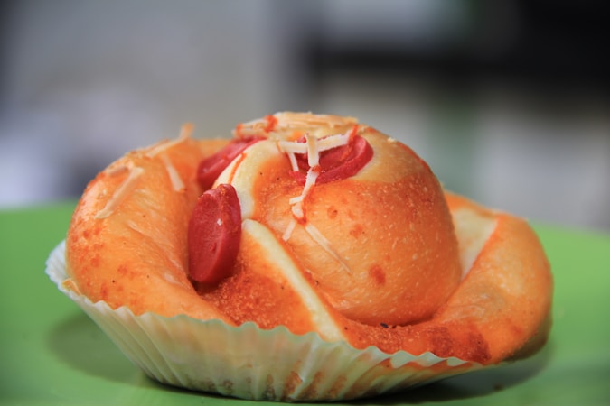 A savory pastry with a golden brown crust, topped with slices of red sausage and sprinkled with grated cheese, placed on a green surface.