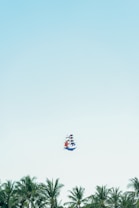 A colorful ship-shaped kite floats in a clear blue sky above a line of lush green palm trees. The kite features red, blue, and white segments, mimicking the appearance of a sailing ship.