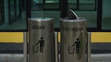 Two metallic trash bins are labeled 'Organik' and 'Non Organik,' standing side by side. The bins have a sleek design with a shiny surface, and each displays a silhouette of a person disposing of trash. The lid of the 'Non Organik' bin is slightly open. They are placed in front of a glass window with a yellow and black backdrop.