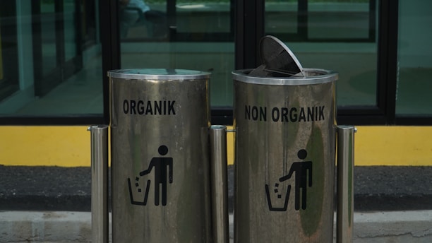 Two metallic trash bins are labeled 'Organik' and 'Non Organik,' standing side by side. The bins have a sleek design with a shiny surface, and each displays a silhouette of a person disposing of trash. The lid of the 'Non Organik' bin is slightly open. They are placed in front of a glass window with a yellow and black backdrop.