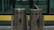 Two metallic trash bins are labeled 'Organik' and 'Non Organik,' standing side by side. The bins have a sleek design with a shiny surface, and each displays a silhouette of a person disposing of trash. The lid of the 'Non Organik' bin is slightly open. They are placed in front of a glass window with a yellow and black backdrop.