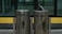 Two metallic trash bins are labeled 'Organik' and 'Non Organik,' standing side by side. The bins have a sleek design with a shiny surface, and each displays a silhouette of a person disposing of trash. The lid of the 'Non Organik' bin is slightly open. They are placed in front of a glass window with a yellow and black backdrop.
