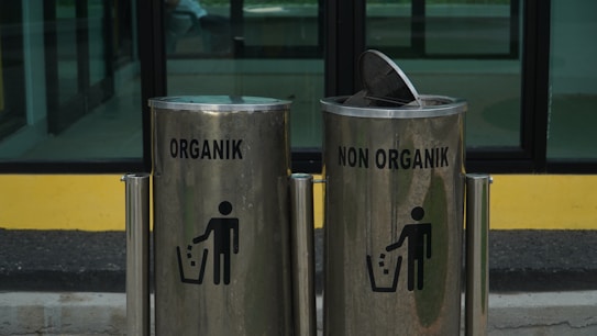 Two metallic trash bins are labeled 'Organik' and 'Non Organik,' standing side by side. The bins have a sleek design with a shiny surface, and each displays a silhouette of a person disposing of trash. The lid of the 'Non Organik' bin is slightly open. They are placed in front of a glass window with a yellow and black backdrop.