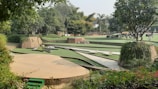 A landscaped garden featuring various levels of green lawns bordered by brick walls. Trees and lush greenery surround the area, providing a serene and natural setting. There are paved paths and some seating areas integrated into the design.