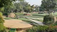 A landscaped garden featuring various levels of green lawns bordered by brick walls. Trees and lush greenery surround the area, providing a serene and natural setting. There are paved paths and some seating areas integrated into the design.