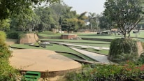A landscaped garden featuring various levels of green lawns bordered by brick walls. Trees and lush greenery surround the area, providing a serene and natural setting. There are paved paths and some seating areas integrated into the design.