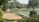 A landscaped garden featuring various levels of green lawns bordered by brick walls. Trees and lush greenery surround the area, providing a serene and natural setting. There are paved paths and some seating areas integrated into the design.