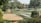 A landscaped garden featuring various levels of green lawns bordered by brick walls. Trees and lush greenery surround the area, providing a serene and natural setting. There are paved paths and some seating areas integrated into the design.