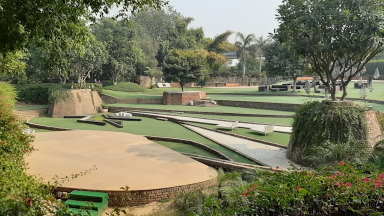 A landscaped garden featuring various levels of green lawns bordered by brick walls. Trees and lush greenery surround the area, providing a serene and natural setting. There are paved paths and some seating areas integrated into the design.