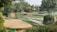 A landscaped garden featuring various levels of green lawns bordered by brick walls. Trees and lush greenery surround the area, providing a serene and natural setting. There are paved paths and some seating areas integrated into the design.