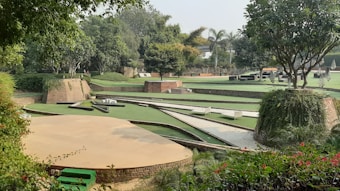 A landscaped garden featuring various levels of green lawns bordered by brick walls. Trees and lush greenery surround the area, providing a serene and natural setting. There are paved paths and some seating areas integrated into the design.