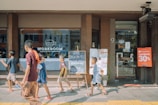 Several children are walking in front of a coffee shop called 'WORKROOM Coffee & Collaboration.' A blackboard outside the shop advertises a 15% discount, and a sign nearby offers a 30% cashback promotion. The environment appears busy and lively, with reflections in the large glass window of the shop.