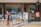 Several children are walking in front of a coffee shop called 'WORKROOM Coffee & Collaboration.' A blackboard outside the shop advertises a 15% discount, and a sign nearby offers a 30% cashback promotion. The environment appears busy and lively, with reflections in the large glass window of the shop.