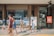 Several children are walking in front of a coffee shop called 'WORKROOM Coffee & Collaboration.' A blackboard outside the shop advertises a 15% discount, and a sign nearby offers a 30% cashback promotion. The environment appears busy and lively, with reflections in the large glass window of the shop.
