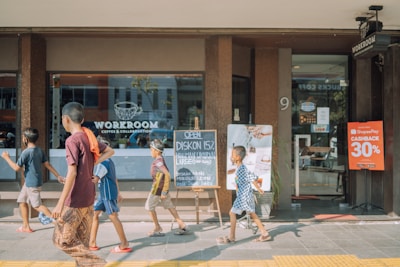 Several children are walking in front of a coffee shop called 'WORKROOM Coffee & Collaboration.' A blackboard outside the shop advertises a 15% discount, and a sign nearby offers a 30% cashback promotion. The environment appears busy and lively, with reflections in the large glass window of the shop.