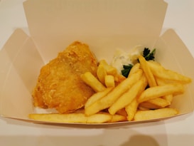 A serving of golden-brown battered fish and crispy fries is placed in a plain, open cardboard box. There is a small amount of creamy tartar sauce and a sprig of parsley as garnish beside the fish.