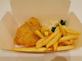 A serving of golden-brown battered fish and crispy fries is placed in a plain, open cardboard box. There is a small amount of creamy tartar sauce and a sprig of parsley as garnish beside the fish.