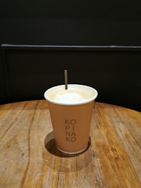 A paper cup filled with coffee sits on a wooden table. The cup has 'KOPI NA KO' printed on it and a small stirrer sticks out from the top. The background is dark, giving a contrast to the light-colored cup and table.