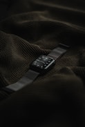 A smart watch with a dark screen and metallic strap is placed on top of a textured dark green fabric.