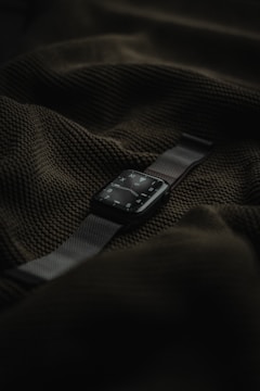 A smart watch with a dark screen and metallic strap is placed on top of a textured dark green fabric.