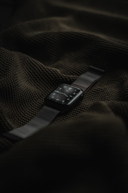 A smart watch with a dark screen and metallic strap is placed on top of a textured dark green fabric.