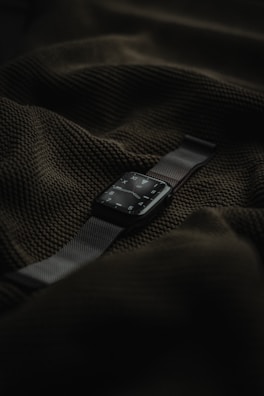 A smart watch with a dark screen and metallic strap is placed on top of a textured dark green fabric.