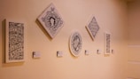 A series of five framed artworks with intricate designs and text are displayed on a light-colored wall. The pieces vary in shape, including rectangular, square, and oval, and each has accompanying labels beneath them.
