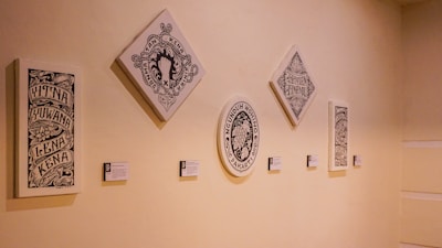 A series of five framed artworks with intricate designs and text are displayed on a light-colored wall. The pieces vary in shape, including rectangular, square, and oval, and each has accompanying labels beneath them.