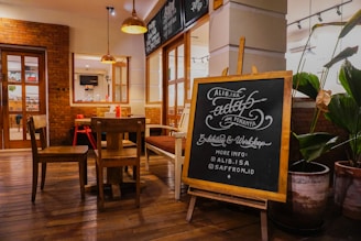 A cozy interior of a café or restaurant with wooden floors and brick walls, featuring a wooden table and chairs. A chalkboard stands prominently, displaying handwritten text about an exhibition and workshop, with social media information. Potted plants decorate the surroundings, adding a touch of greenery.