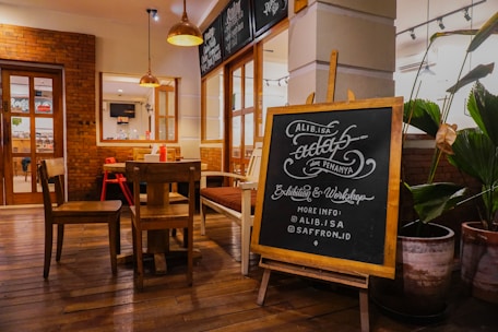 A cozy interior of a café or restaurant with wooden floors and brick walls, featuring a wooden table and chairs. A chalkboard stands prominently, displaying handwritten text about an exhibition and workshop, with social media information. Potted plants decorate the surroundings, adding a touch of greenery.