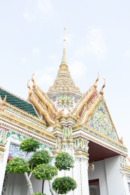An ornate temple structure featuring intricate golden spires and detailed architectural elements. The building is adorned with colorful mosaics and traditional artistic decorations, characteristic of Southeast Asian religious structures. In the foreground, there are lush green trees, enhancing the serene atmosphere.