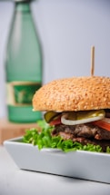 A gourmet hamburger with a sesame seed bun is placed on a rectangular white plate. The burger is garnished with fresh lettuce, slices of tomato, onion, and pickles. In the background, there is a green glass bottle with a label, slightly out of focus.