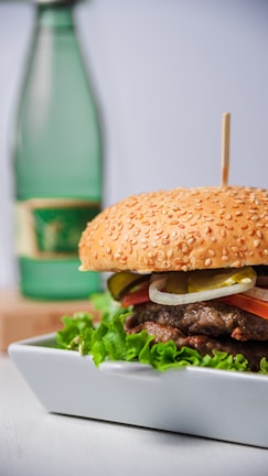 A gourmet hamburger with a sesame seed bun is placed on a rectangular white plate. The burger is garnished with fresh lettuce, slices of tomato, onion, and pickles. In the background, there is a green glass bottle with a label, slightly out of focus.